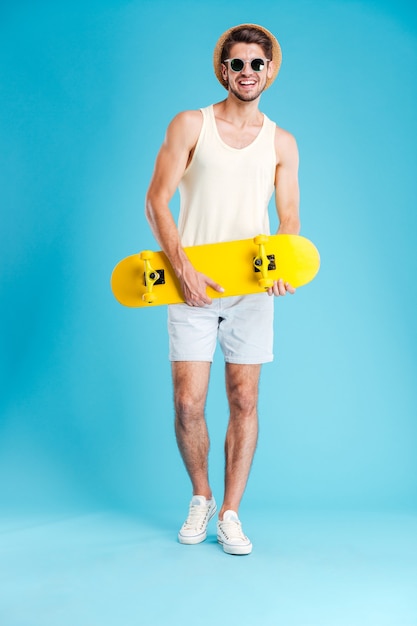 Cheerful young man in hat and sunglasses standing and holding skateboard over blue wall