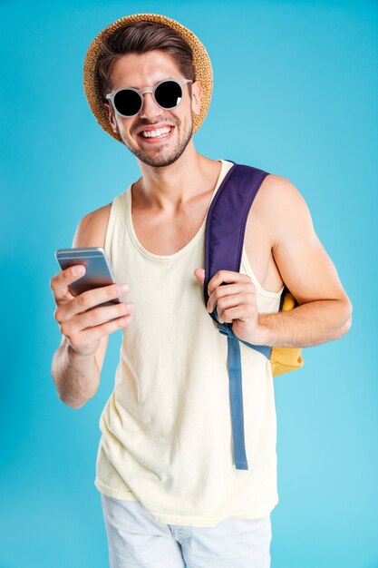 Cheerful young man in hat and sunglasses with backpack using smartphone