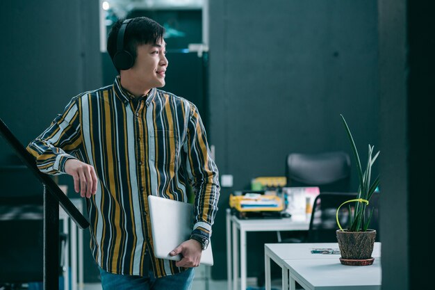 Cheerful young man holding a laptop and looking away while listening to music in headphones