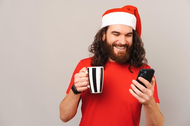 Cheerful young man is holding a hot mug while checking his phone at Christmas