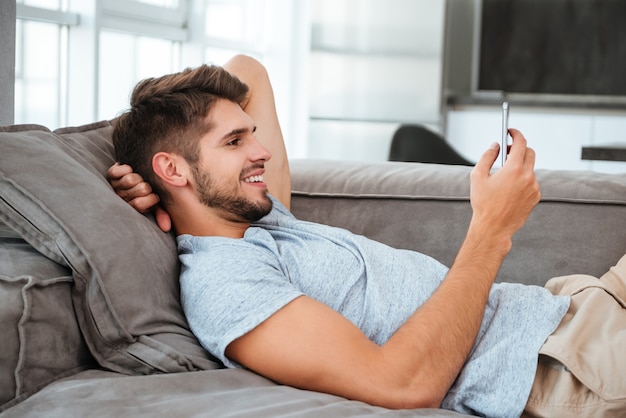 Cheerful young man lies on sofa and looking on phone.