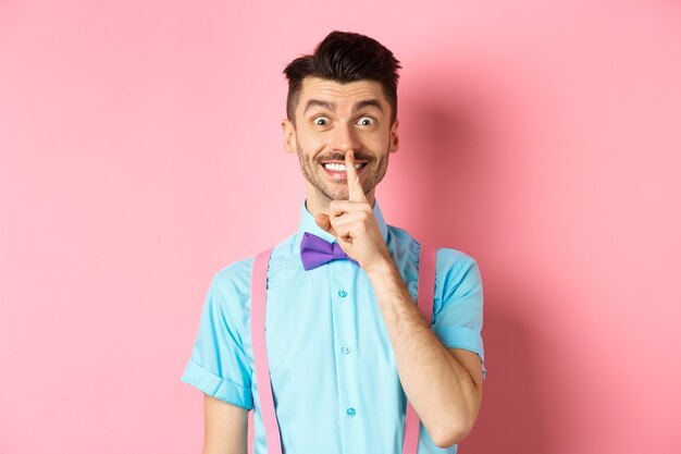 Cheerful young man making surprise, shushing at camera with happy smile, asking to keep voice down, be quiet, standing on pink.