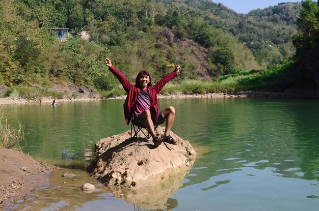 A cheerful young man in a red jacket sits relaxed on the bank of a still beautiful river