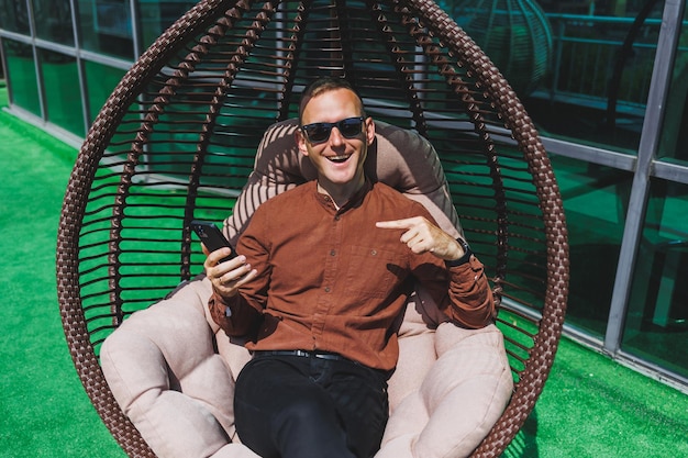 A cheerful young man in a shirt with a phone sits on the terrace of an office building after work uses wireless internet a positive male employee in glasses is talking on the phone