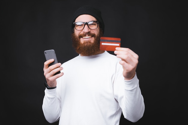 陽気な若い男が彼の新しいクレジットカードを示し、スマートフォンを保持