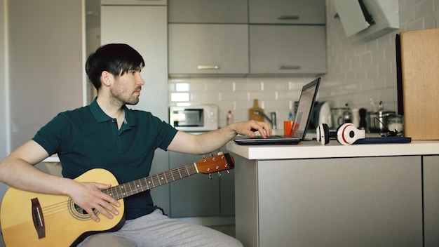 Cheerful young man sitting at kitchen learning to play guitar using laptop computer at home