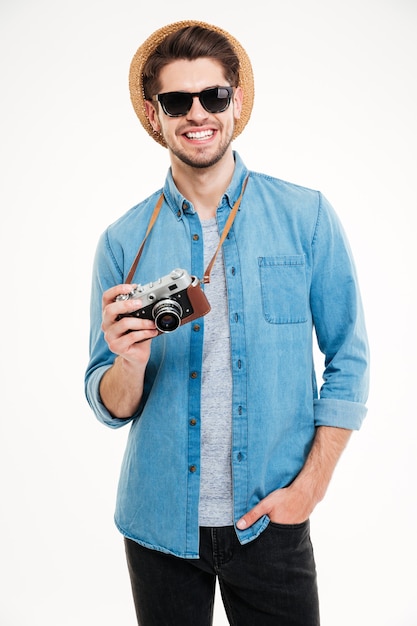 Cheerful young man in sunglasses and hat using old vintage photo camera over white background