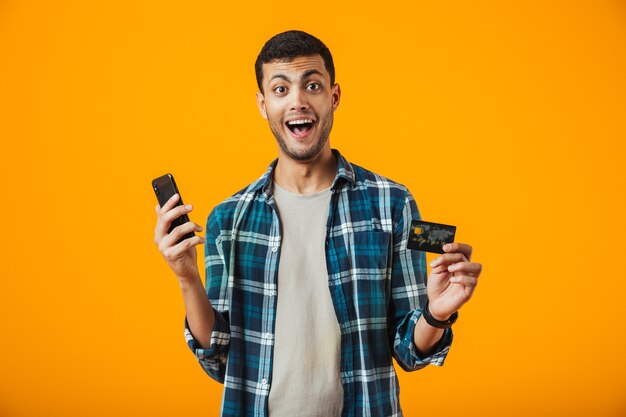 Cheerful young man wearing plaid shirt standing isolated over orange background, holding mobile phone, showing plastic credit card