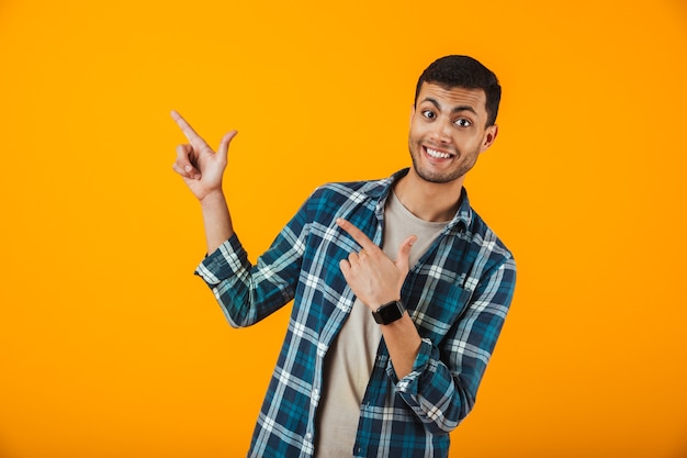 Веселый молодой человек в клетчатой рубашке стоит изолированно над оранжевой стеной, указывая пальцем в сторону