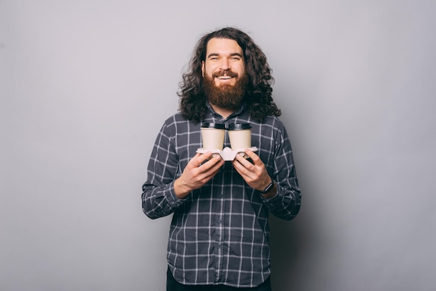 Cheerful young man with beard in checkered shirt holding two cups of coffee to go