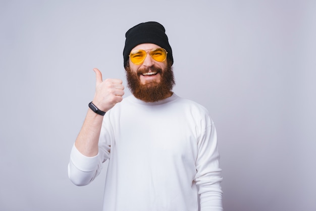 Cheerful young man with beard is smiling at the camera and showing thumb up on white background.