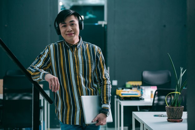 Cheerful young man with headphones on his head smiling while standing with a laptop