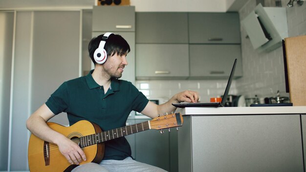 Cheerful young man with headphones sitting at kitchen learning to play guitar using laptop