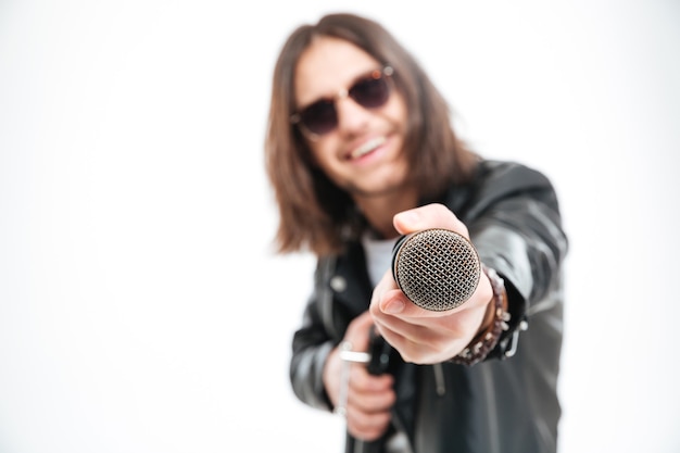 Cheerful young man with long hair in sunglasses giving a microphone and offering you to sing over white background