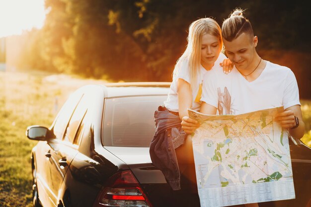 Cheerful young man and woman leaning on trunk of car in nature and reading map