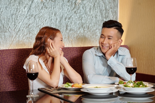 Cheerful young man and woman sitting at restaurant table with tasty dishes and looking at each other