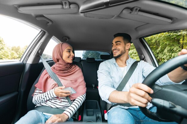 スマートフォンと夫と一緒にヒジャブを着た陽気な中東の若い女性が車に乗る