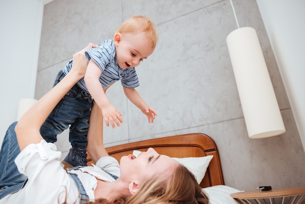 Cheerful young mother laughing and playing with her little son on bed