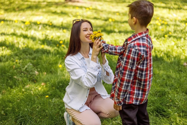 Cheerful young mother smelling flowers from her son mom and kid in park with dandelions