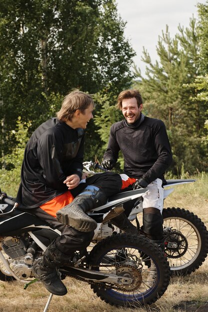 Cheerful young motorcyclists in protective outfits sitting on motorbikes and laughing while chatting in forest