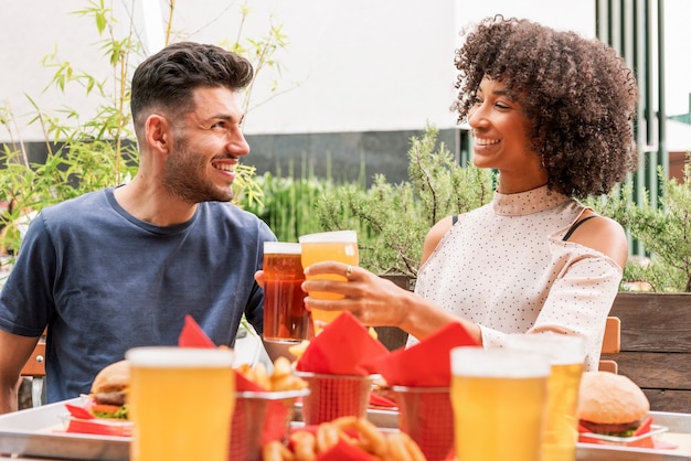 Cheerful young multiracial couple clinking glasses of beer while enjoying summer weekend together in outdoor restaurant and celebrating holiday
