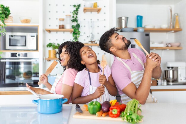 Photo cheerful young parents and little daughter having fun while baking together in kitchen happy african american family singing and fooling using spatula and whisk as microphones copy space