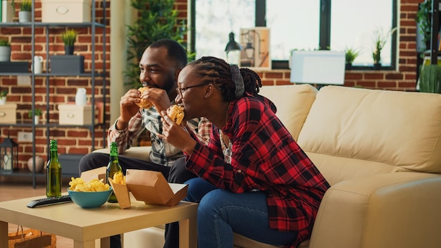 リビングルームでハンバーガーやフライドポテトを食べたり、テレビで好きな映画を見たり、アルコールを飲んだりする陽気な若者。テイクアウトの場所からファーストフードの注文を提供するボーイフレンドとガールフレンド。