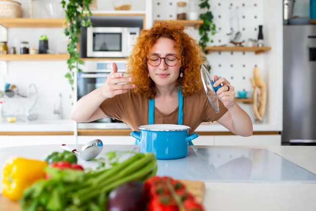 陽気な若い美しい女性は,前を着て,自宅のキッチンで昼食を準備し,料理を嗅いでいます. 女性は,自宅で新しいレシピで家族のために夕食を調理しています.