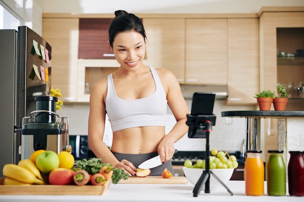 cheerful young pretty sportswoman cooking breakfast in kitchen and recording video for her blog