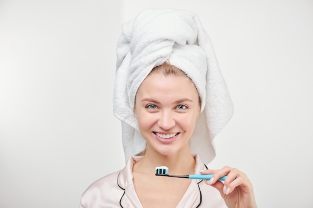 Cheerful young pretty woman with toothy smile holding toothbrush by her mouth while standing in isolation