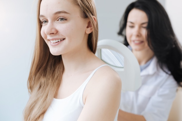 Cheerful young proficient dermatologist working in the clinic and having appointment with patient while using medical loupe for skin examination