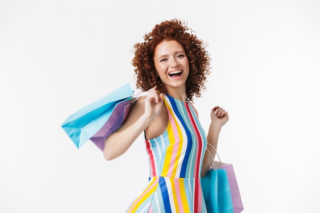 Photo cheerful young redhaired girl wearing dress, carrying shopping bags isolated over white wall