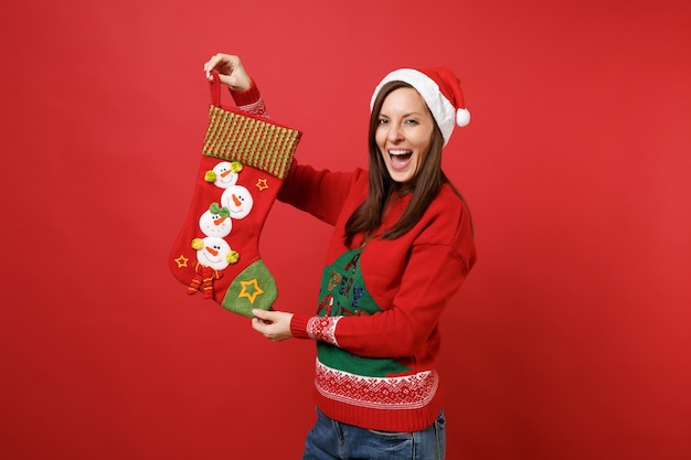 Foto allegra giovane ragazza di babbo natale con il cappello di natale che tiene la bocca spalancata, tiene la calza, calzino per i regali isolato su sfondo rosso. felice anno nuovo 2019 celebrazione festa concetto. mock up copia spazio.