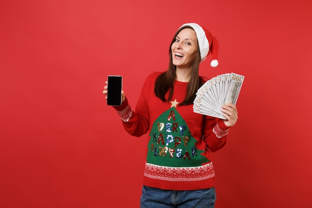 Cheerful young Santa girl holding lots bunch of dollars banknotes cash money and mobile phone with blank empty screen isolated on red background. Happy New Year 2019 celebration holiday party concept.