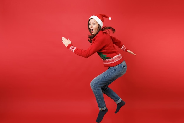 Cheerful young Santa girl in knitted sweater, Christmas hat jumping, keeping mouth wide open isolated on red wall background. Happy New Year 2019 celebration holiday party concept. Mock up copy space.
