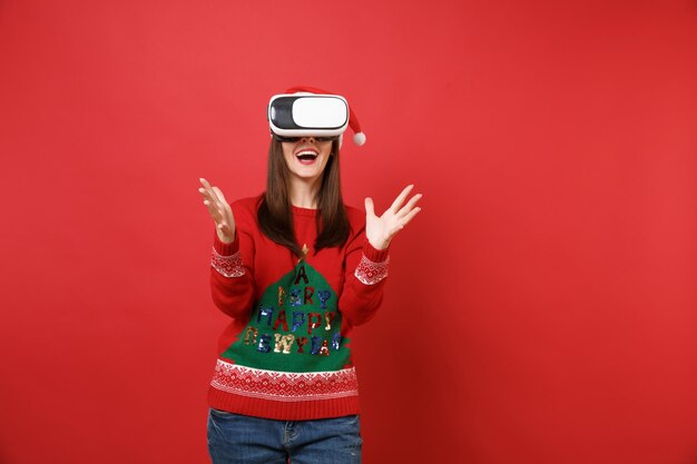 Cheerful young Santa girl in knitted sweater, Christmas hat looking in headset, spreading hands isolated on red background. Happy New Year 2019 celebration holiday party concept. Mock up copy space.