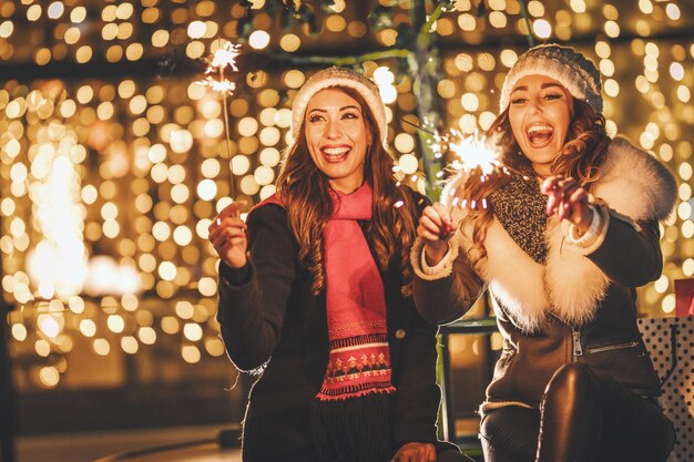 Photo cheerful young sisters are holding sparklers and having fun at holiday winter night in the city street.