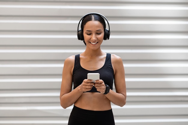 Cheerful young slim arab female in sports uniform and headphones typing on smartphone on gray wall