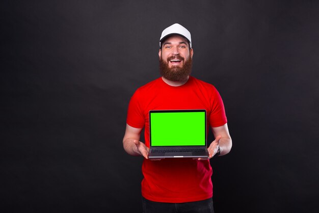 赤いTシャツを着て、ノートパソコンで緑色の画面を表示している陽気な若い笑顔のひげを生やした男