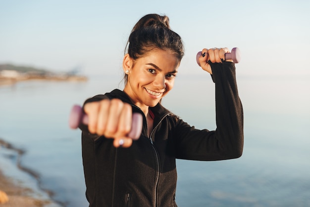 Cheerful young sportsgirl doing exercises with small dumbbells