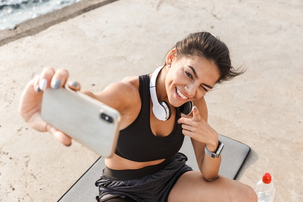 Cheerful young sportswoman resting after workout at the beach, taking a selfie, drinking water, sitting on a fitness mat