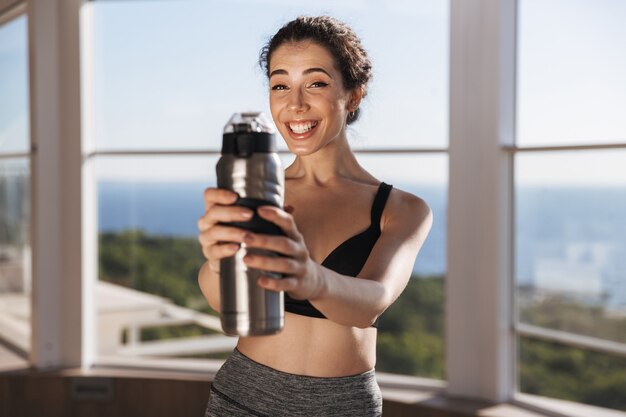 Cheerful young sportswoman showing water bottle