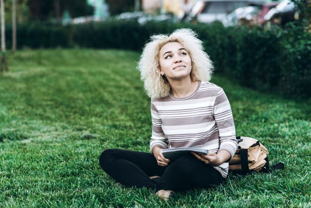 Cheerful young student girl sits on a grass in campus