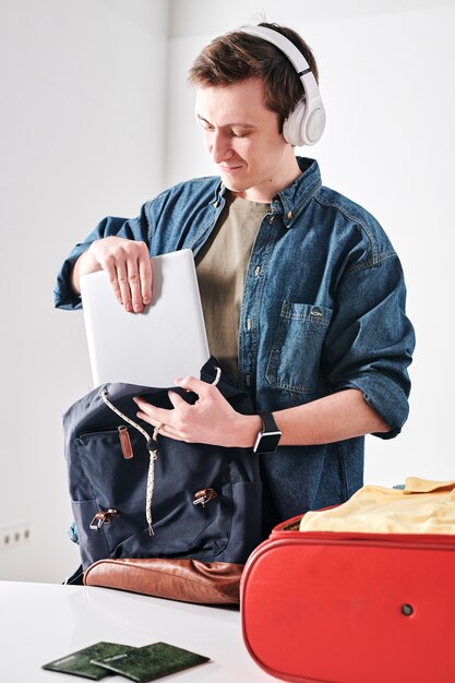 Cheerful young student in wireless headphones packing tablet in satchel while going to travel