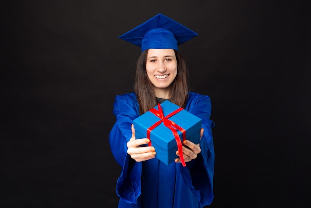 Cheerful young student woman in bachelor and graduating hat holding a gift box