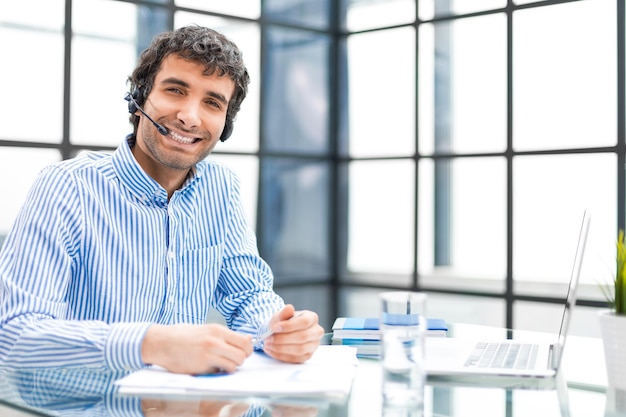 Cheerful young support phone male operator in headset at workplace while using computer help service and client consulting call center concept