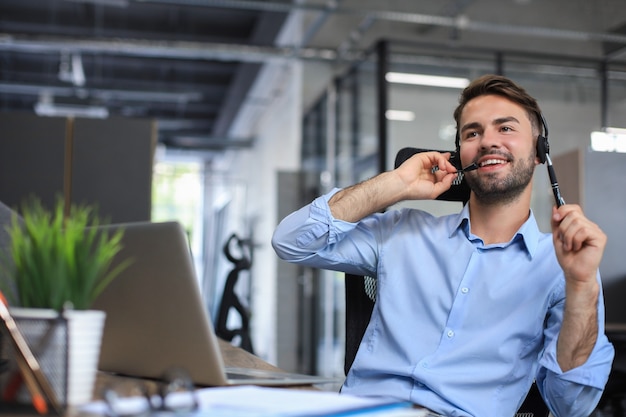 Cheerful young support phone male operator in headset, at workplace while using laptop, help service and client consulting call center concept.