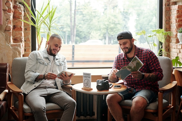 Cheerful young tourists sitting against window and planning travel route using online and paper maps