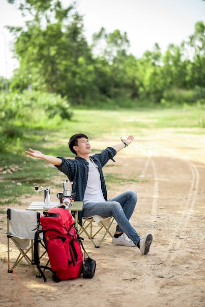 Cheerful Young traveler man sitting at front of the tent in forest with coffee set and making fresh coffee grinder while camping trip on summer vacation