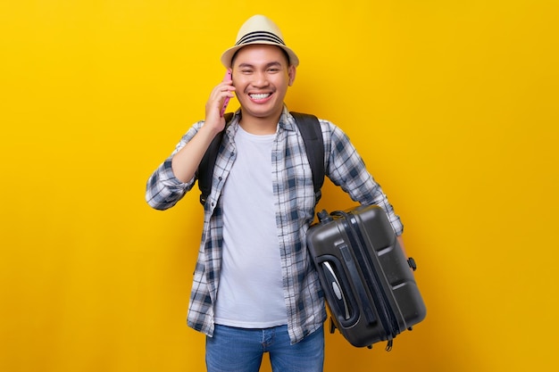 Cheerful young traveler tourist asian man 20 years old wears\
casual clothes hat with backpack holding suitcase and talk speaks\
on mobile phone isolated on yellow background air flight journey\
concept